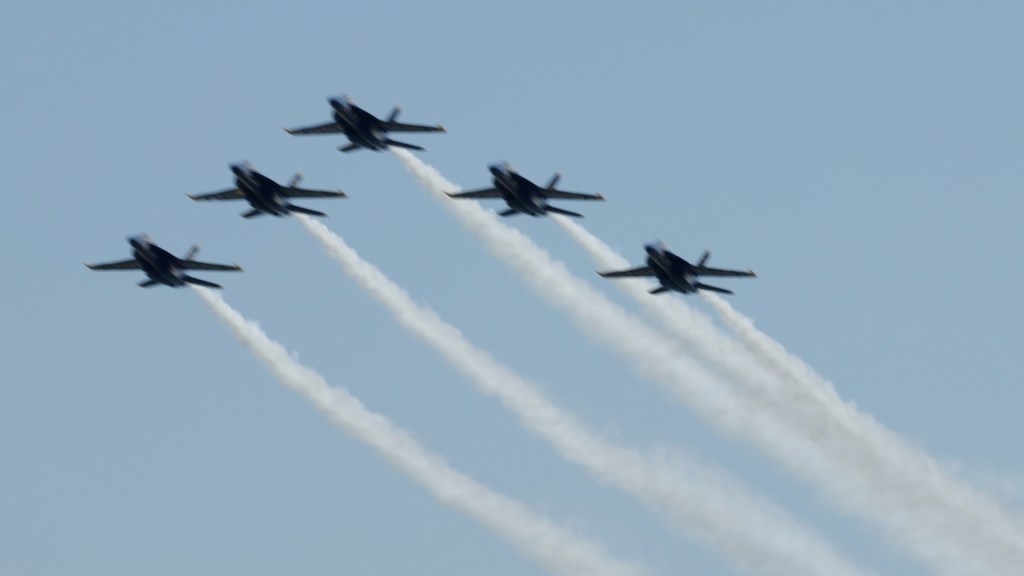 3 Blue Angel jets fly in a triangle formation in the clear blue sky, leaving behind a trail of white smoke. 